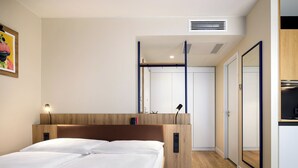 Ein modernes Hotelzimmer mit einem hölzernen Kopfteil, einem Nachttisch mit Lampe, einem großen Spiegel und einem Kleiderschrank.