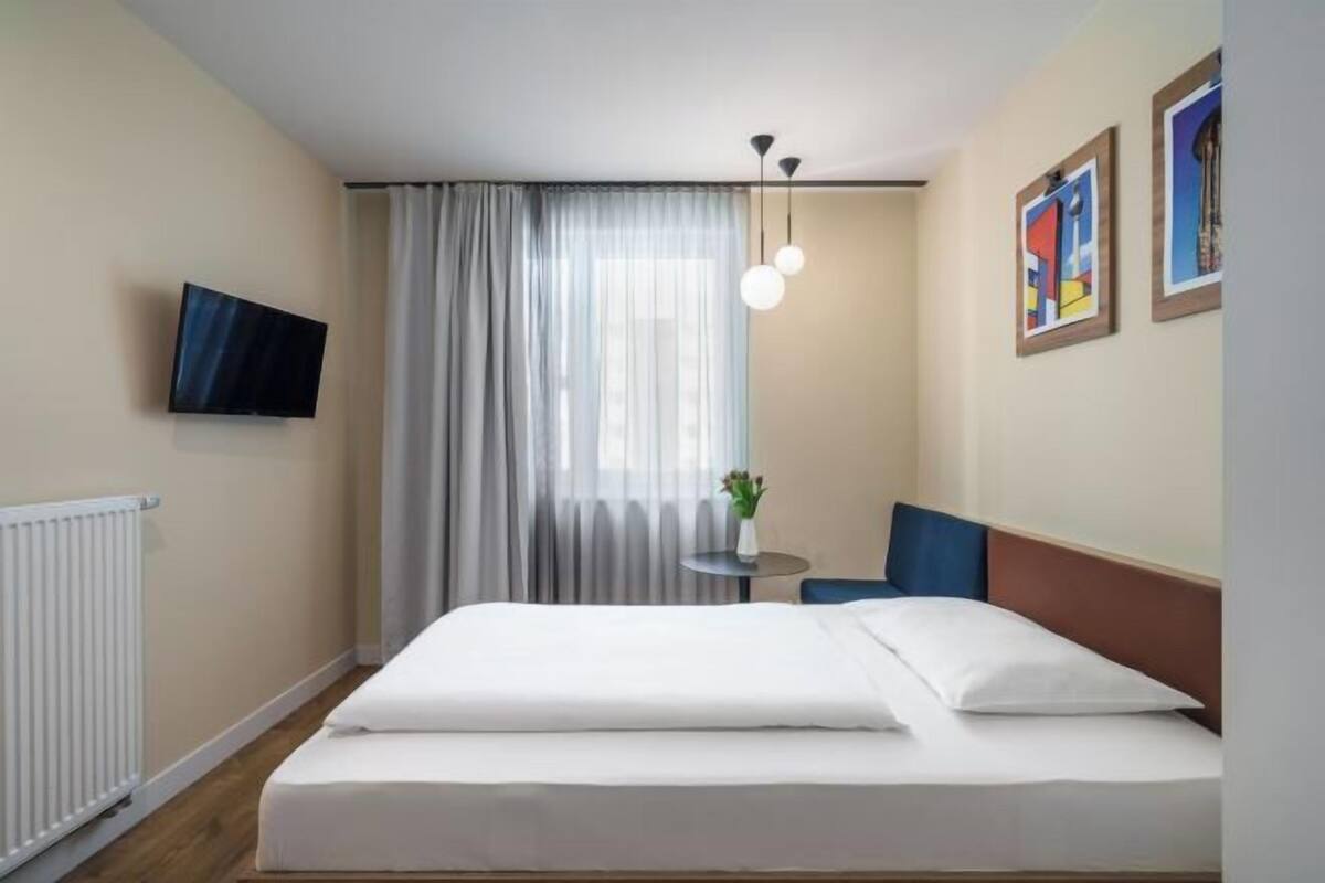 Habitación de hotel con una cama, una mesita con un florero, una silla, un televisor en la pared y una ventana con cortinas.