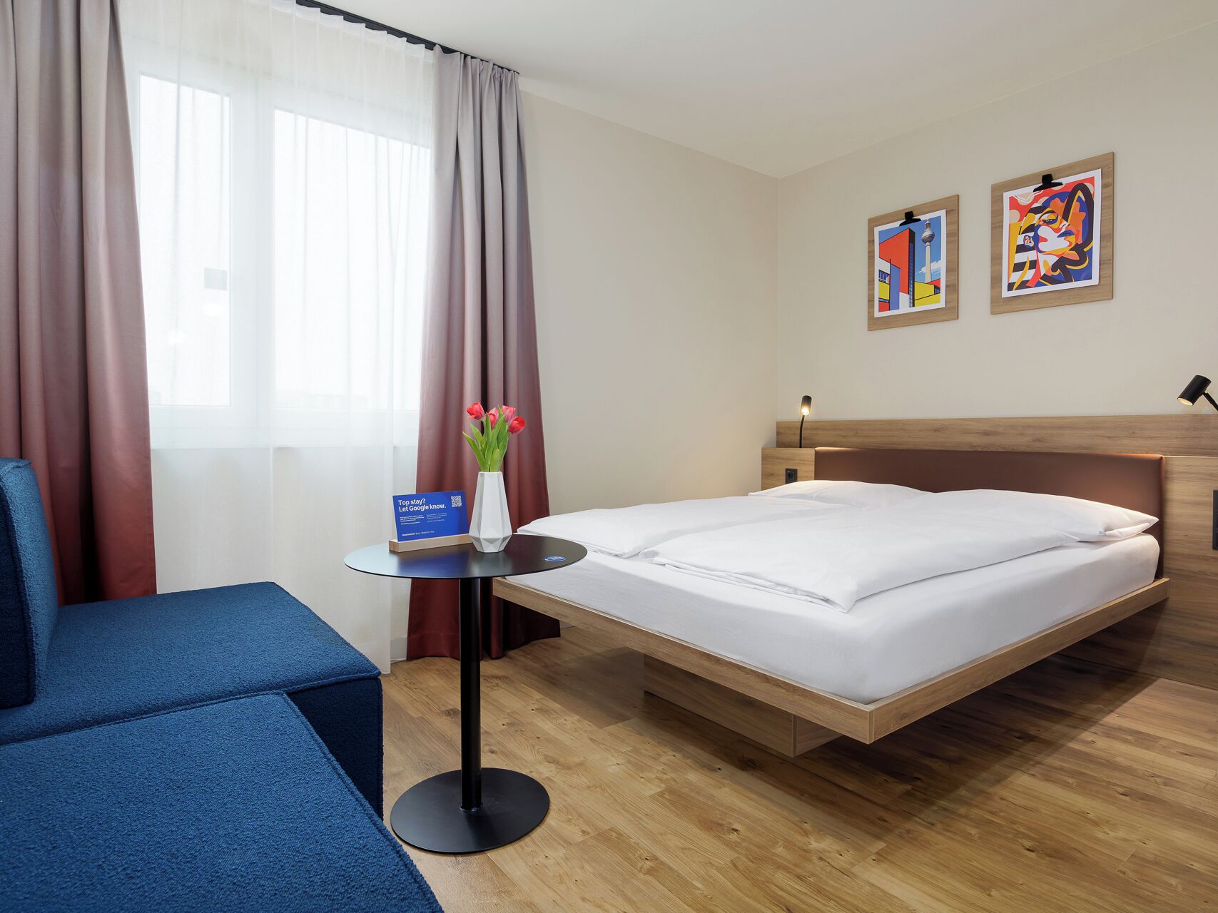 A hotel room with a bed, a small table with a vase of flowers, a blue chair, and two paintings on the wall.
