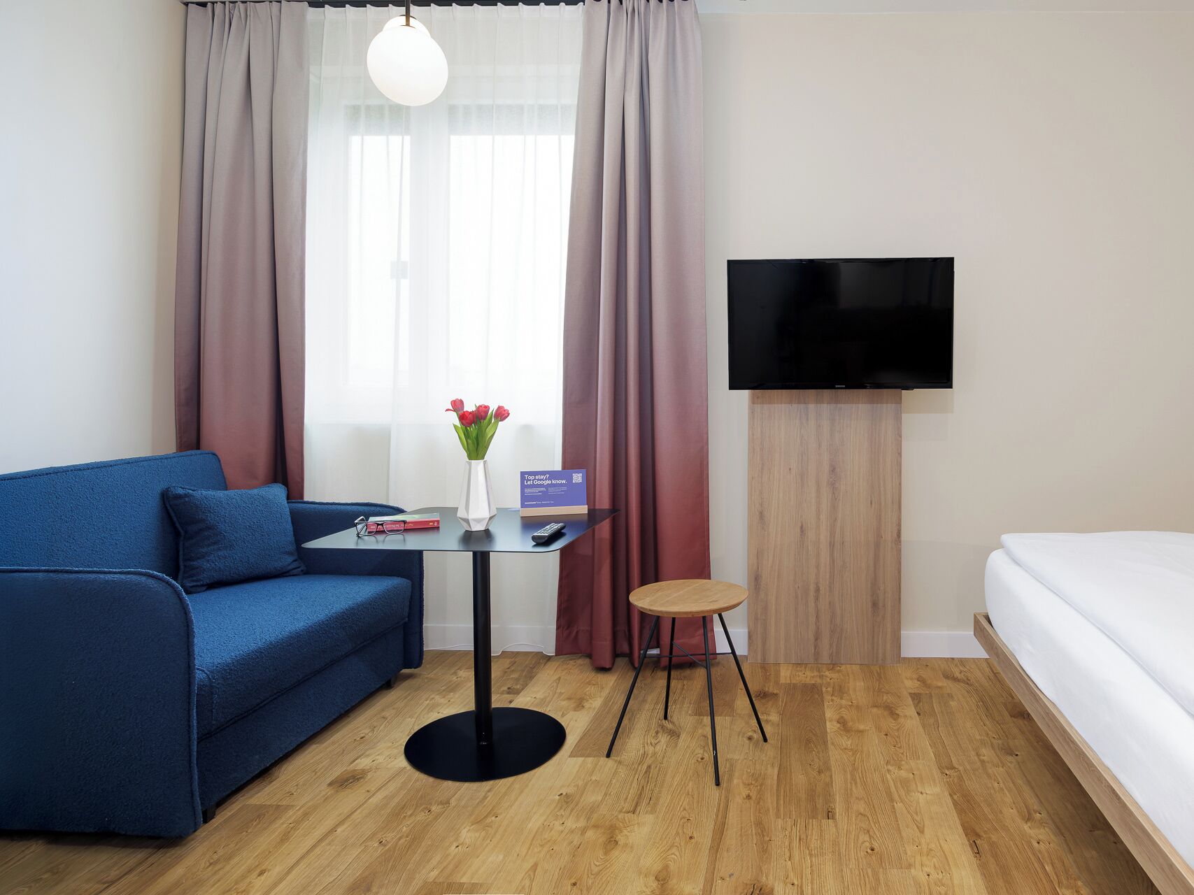 Une chambre d’hôtel avec un canapé bleu, une petite table sur laquelle est posé un vase de fleurs, une télévision et un lit.