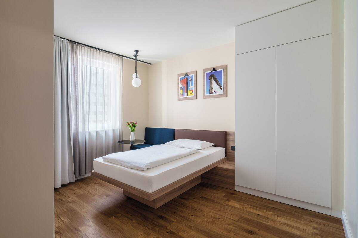 Un dormitorio moderno con piso de madera, una cama con sábanas blancas, una mesita de noche, un florero con flores y dos cuadros en la pared.