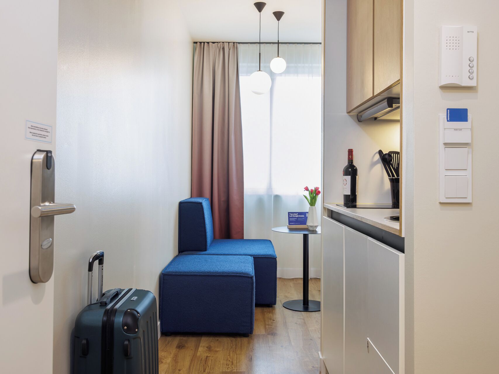 A compact hotel room with a bed, a small kitchenette, a window with curtains, a desk with a bottle of wine, and a suitcase.