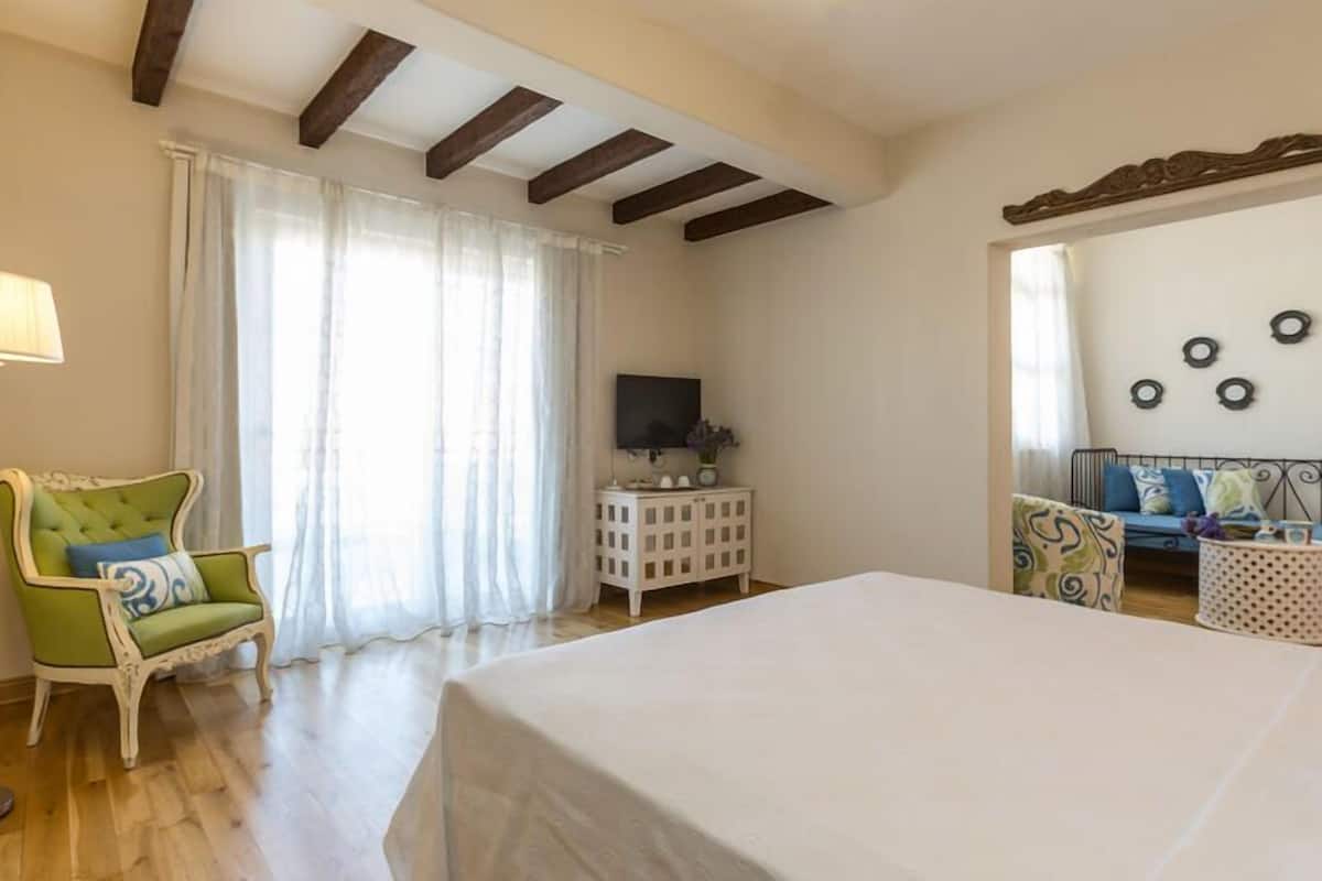 A bedroom with a bed, a green armchair, a small white cabinet, a TV, and a blue sofa.