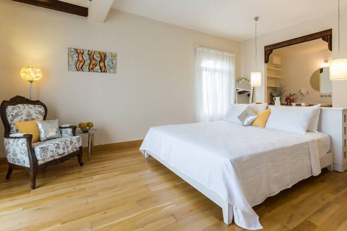 A bedroom with a large bed, a chair with a floral pattern, a small table with a vase, and a painting on the wall.