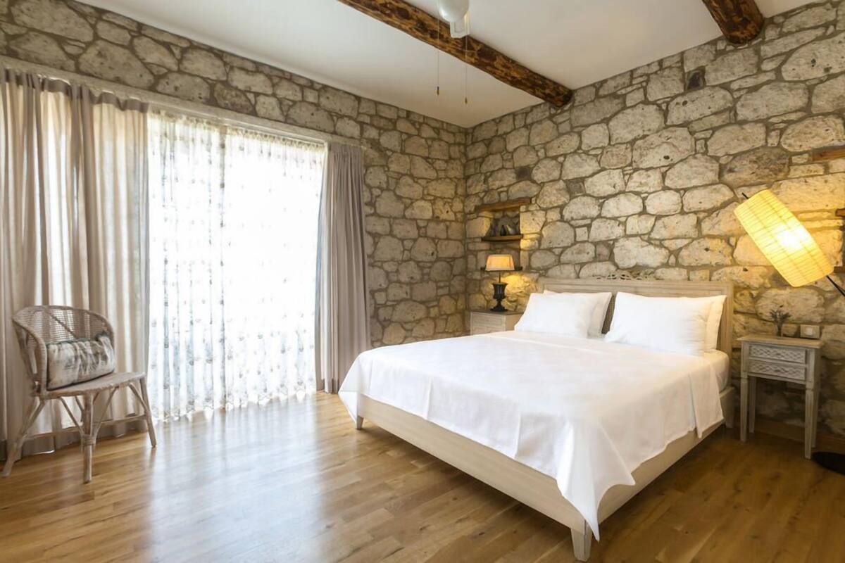 A bedroom with a stone wall, a bed with white linens, a wooden floor, a chair, and a nightstand with a lamp.