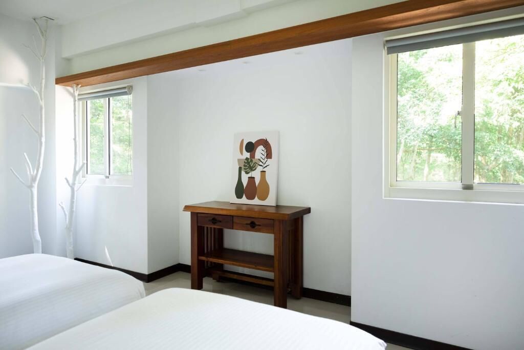 A bedroom with two beds, a wooden nightstand with a vase painting, and large windows with a view of trees.