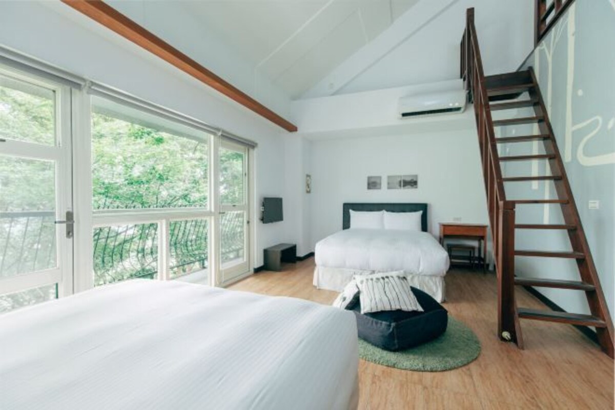 A bedroom with a large bed, a wooden floor, a ladder, and a view through large windows.
