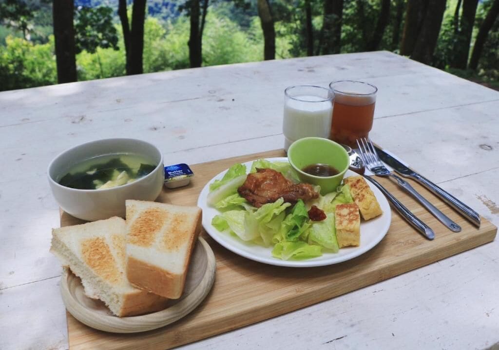 A picnic setup with a plate of food, two glasses of beverages, and a bowl of soup on a wooden table outdoors.