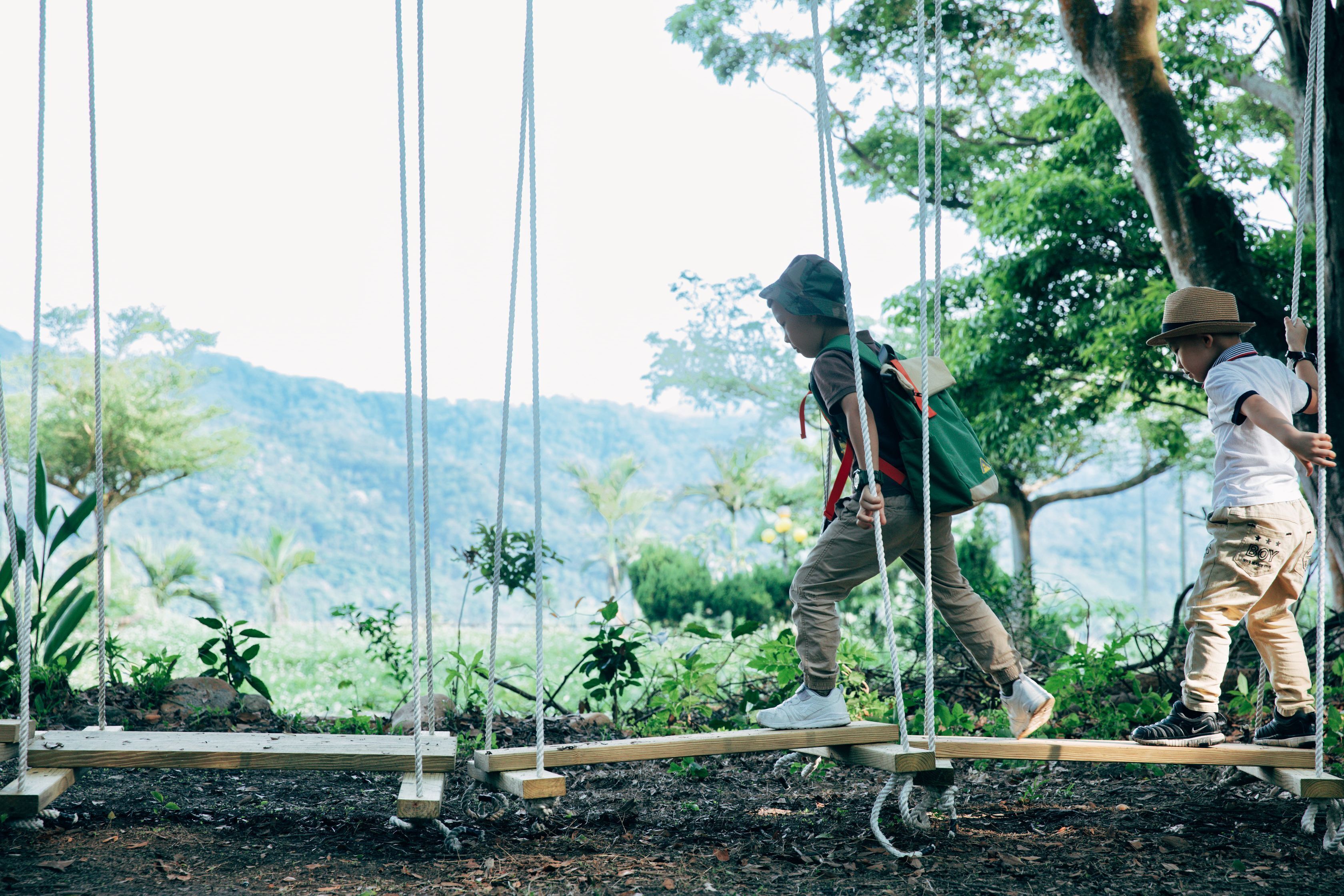 Two children on a rope bridge in a natural setting with trees and a mountainous background.
