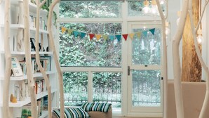 A cozy reading nook with a bookshelf, a cushioned bench, and a view of greenery through large windows. - Lvbolang by REINDEER, Sanyi (Sanyi)