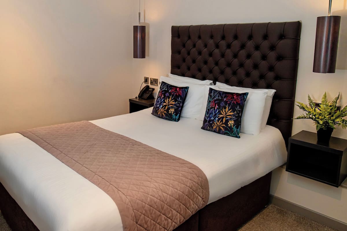 Una cama bien hecha con colcha marrón, cabecera capitonada y almohadas decorativas. Hay dos mesitas de noche con lámparas y una planta.