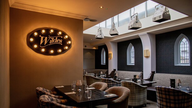 Un restaurant avec une enseigne portant le nom « Wildes », des chaises à carreaux et un plafond haut orné de luminaires suspendus.