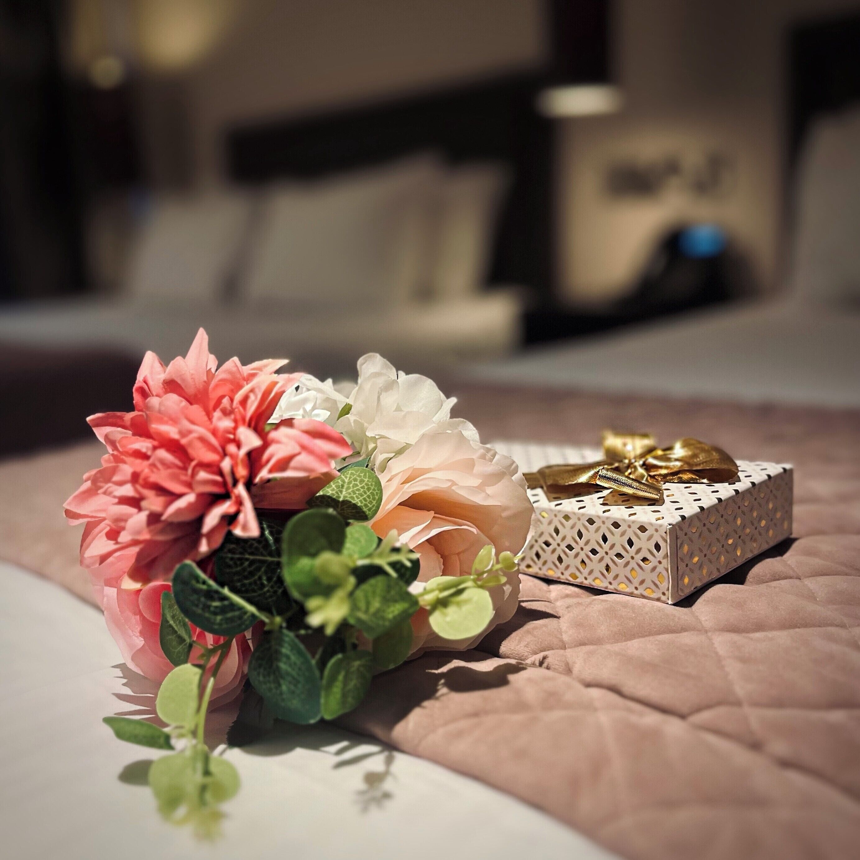 Una cama con un edredón rosa, una caja decorativa blanca con lazo dorado y un ramo de flores rosa y blanca.