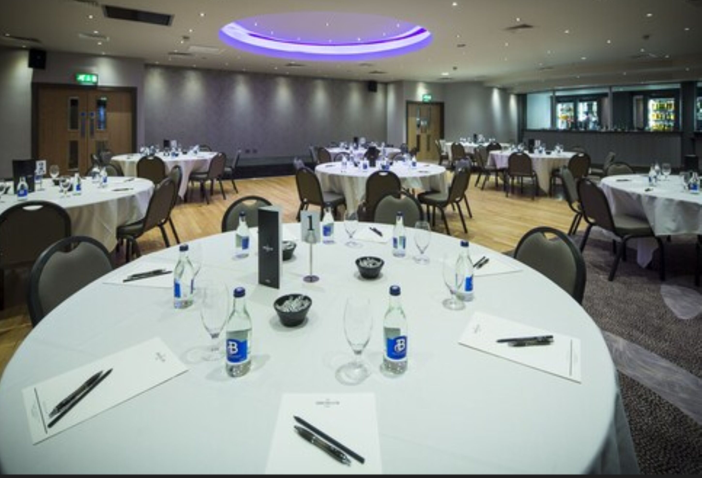 A conference room with round tables, each set with water bottles, glasses, and notepads.