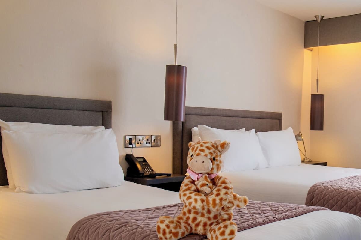 Habitación de hotel con dos camas, una mesita de noche con teléfono y un peluche de jirafa sobre la cama.