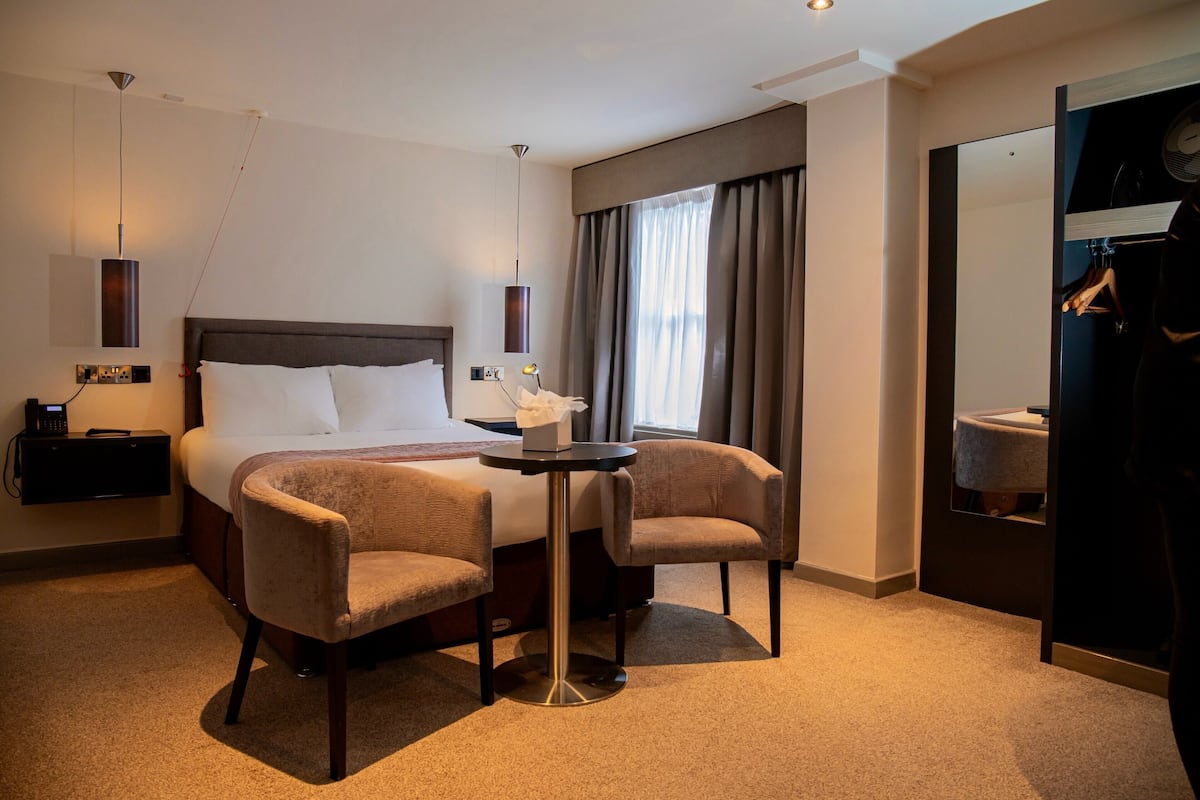 Habitación de hotel con una cama grande, dos sillones, una mesita, una mesita de noche con lámpara, un espejo y una ventana con cortinas.