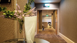 Un hall d’hôtel avec un sol recouvert de moquette, un comptoir d’accueil et une enseigne fixée au mur sur laquelle on peut lire : « L’ACCUEIL LE PLUS CHALEUREUX ».