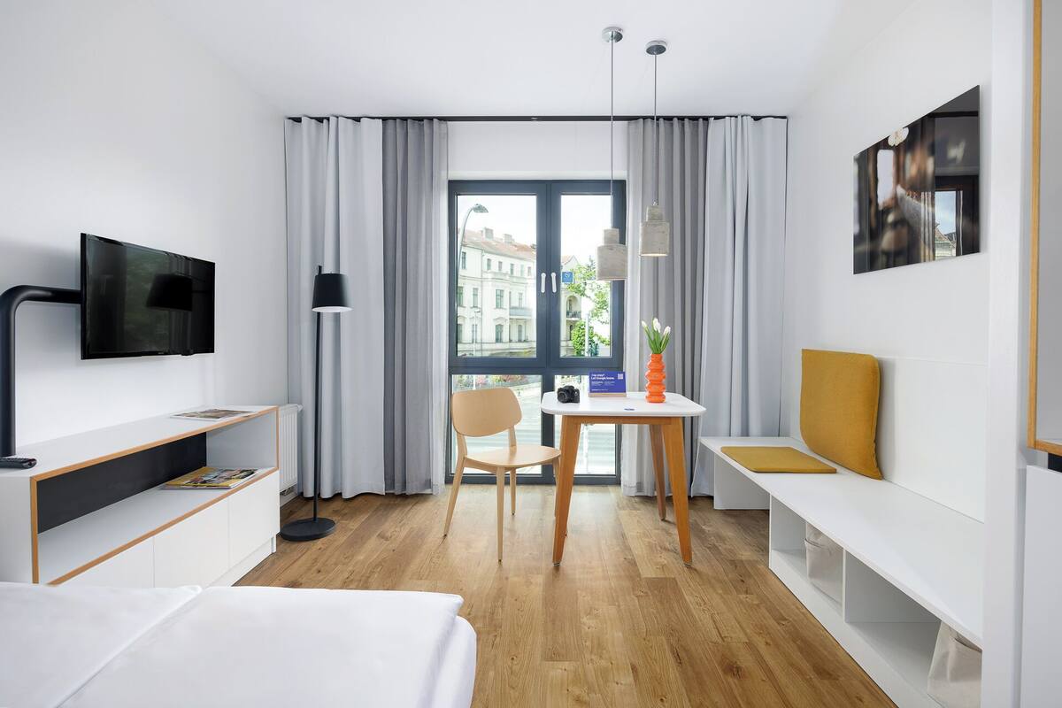 A hotel room with a bed, a desk, a chair, a TV, a lamp, a window with curtains, and a view of buildings outside.