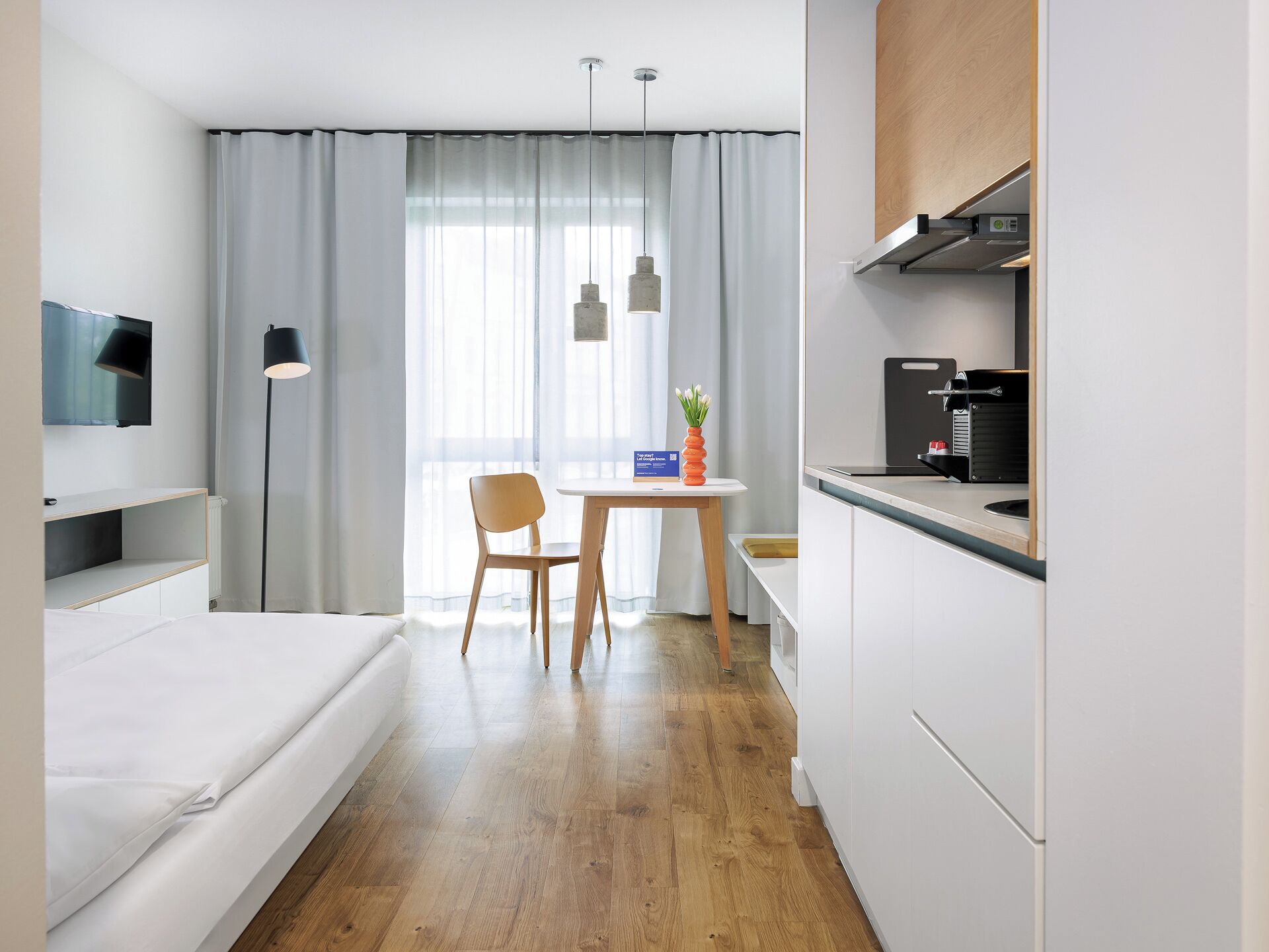 A modern hotel room with a bed, a desk, a chair, a dining table, and a kitchenette.