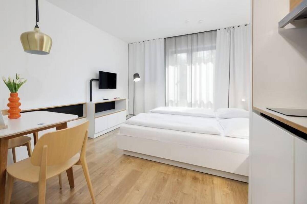 A modern bedroom with a bed, a desk, a chair, a TV, and a window with curtains.