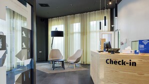 Un mostrador de recepción con detalles de madera, un área de espera con dos sillas, un ventanal con cortinas y un cartel que indica WiFi gratuito.