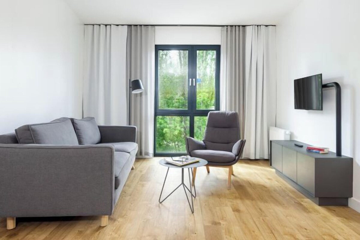 A modern living room with a grey sofa, a grey armchair, a small round table, a TV on the wall, and a window with curtains.