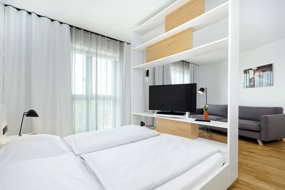 A modern hotel room with a bed, a TV, a sofa, and a desk with a lamp.