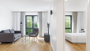 Una sala moderna con un sofá y un sillón grises, una mesita y un mueble para televisión. Hay ventanas grandes con cortinas, piso de madera y una pared blanca.