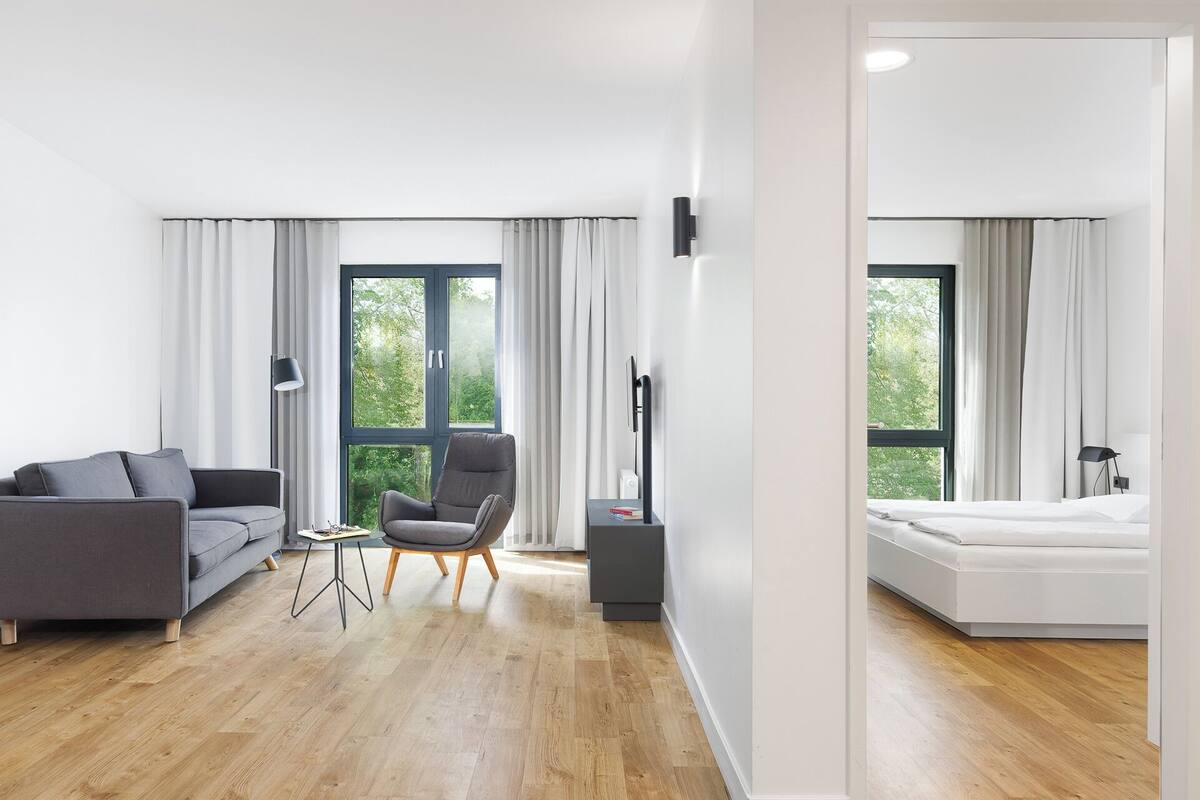 A modern living room with a grey sofa, a grey armchair, a small table, and a TV stand. There are large windows with curtains, a wooden floor, and a white wall.