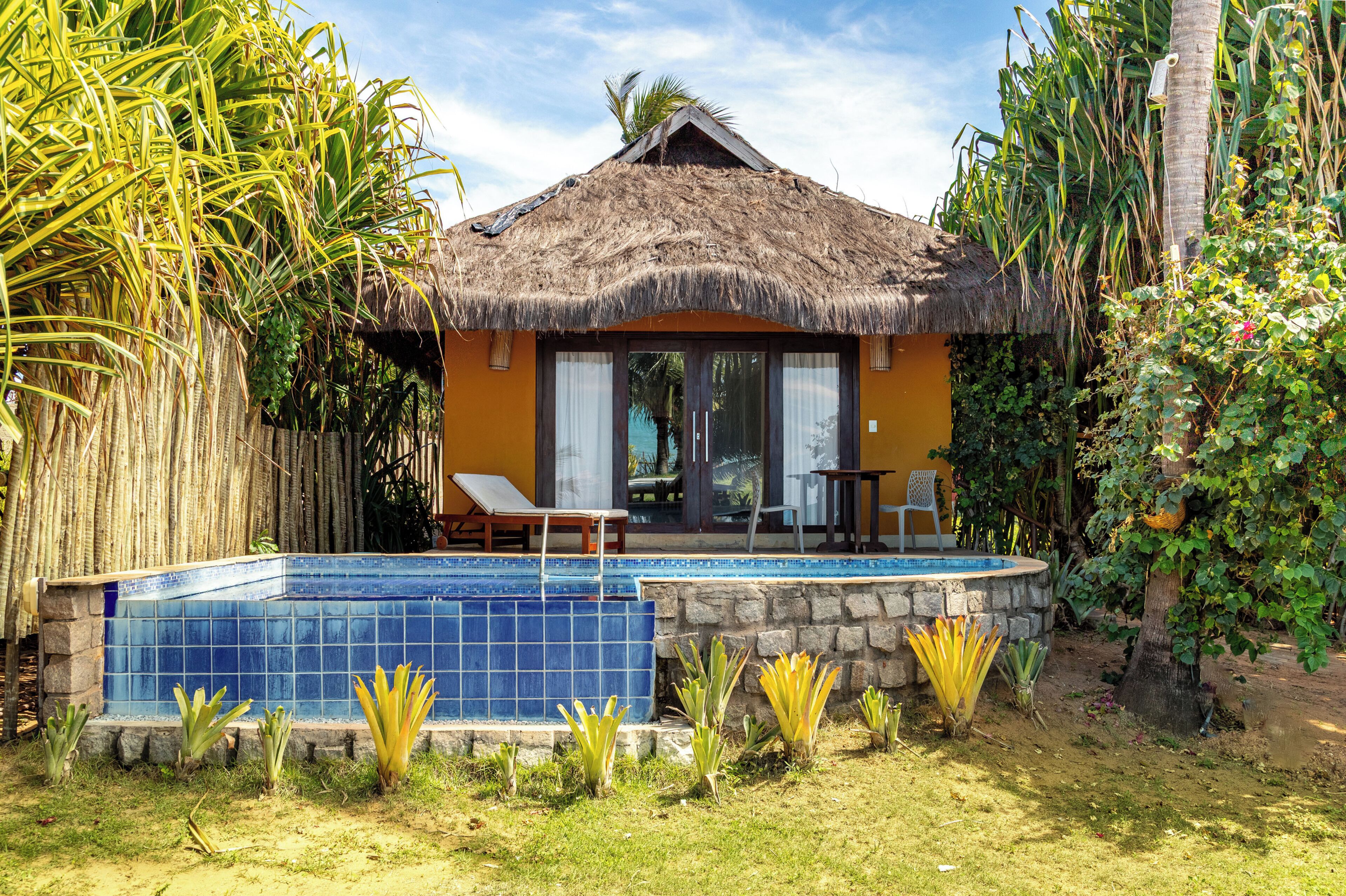 Un bungalow au toit de chaume, doté d’une piscine et entouré de plantes tropicales.
