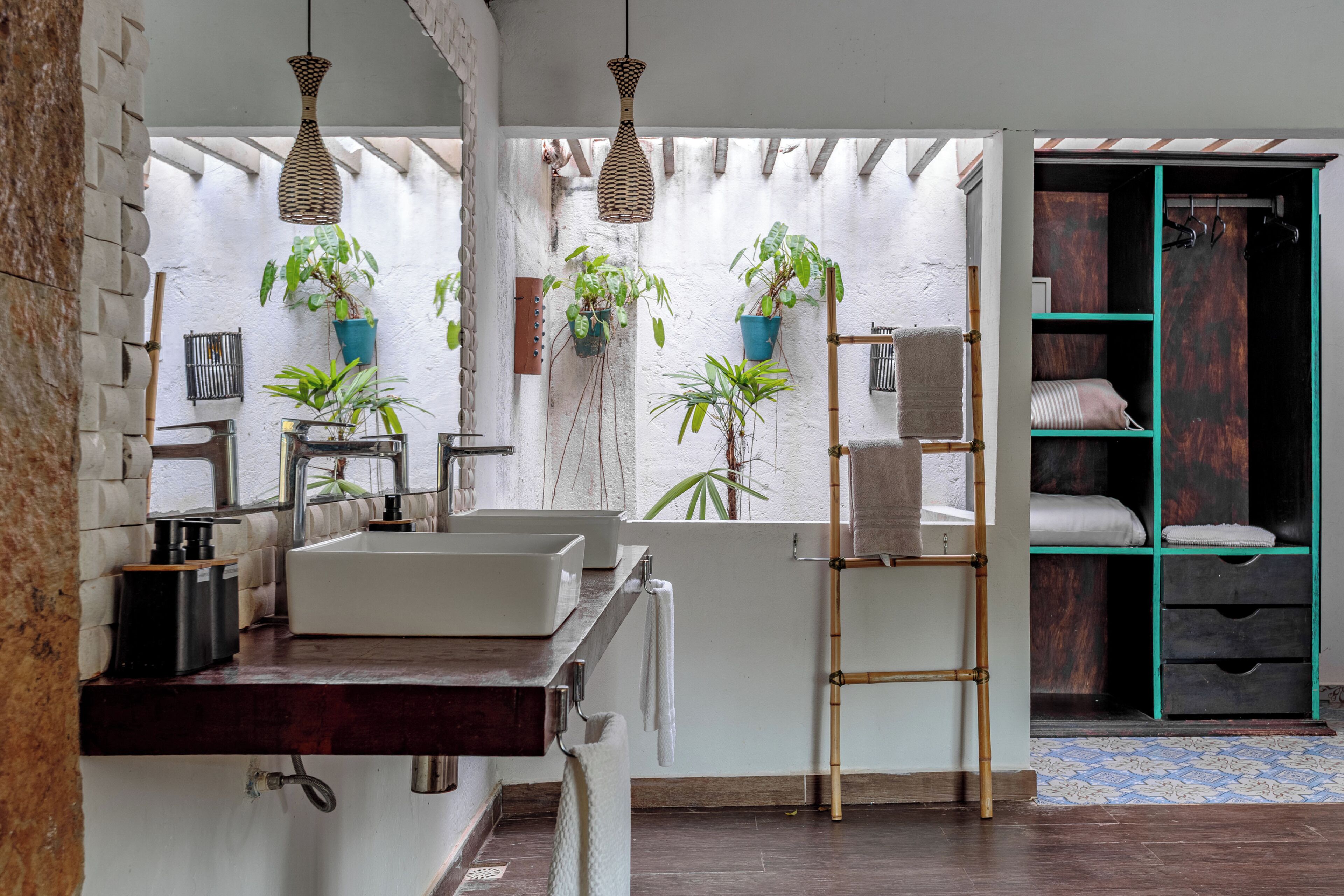 A bathroom with two sinks, a mirror, a towel rack, and a shelf with towels. There are potted plants and a wooden ladder.