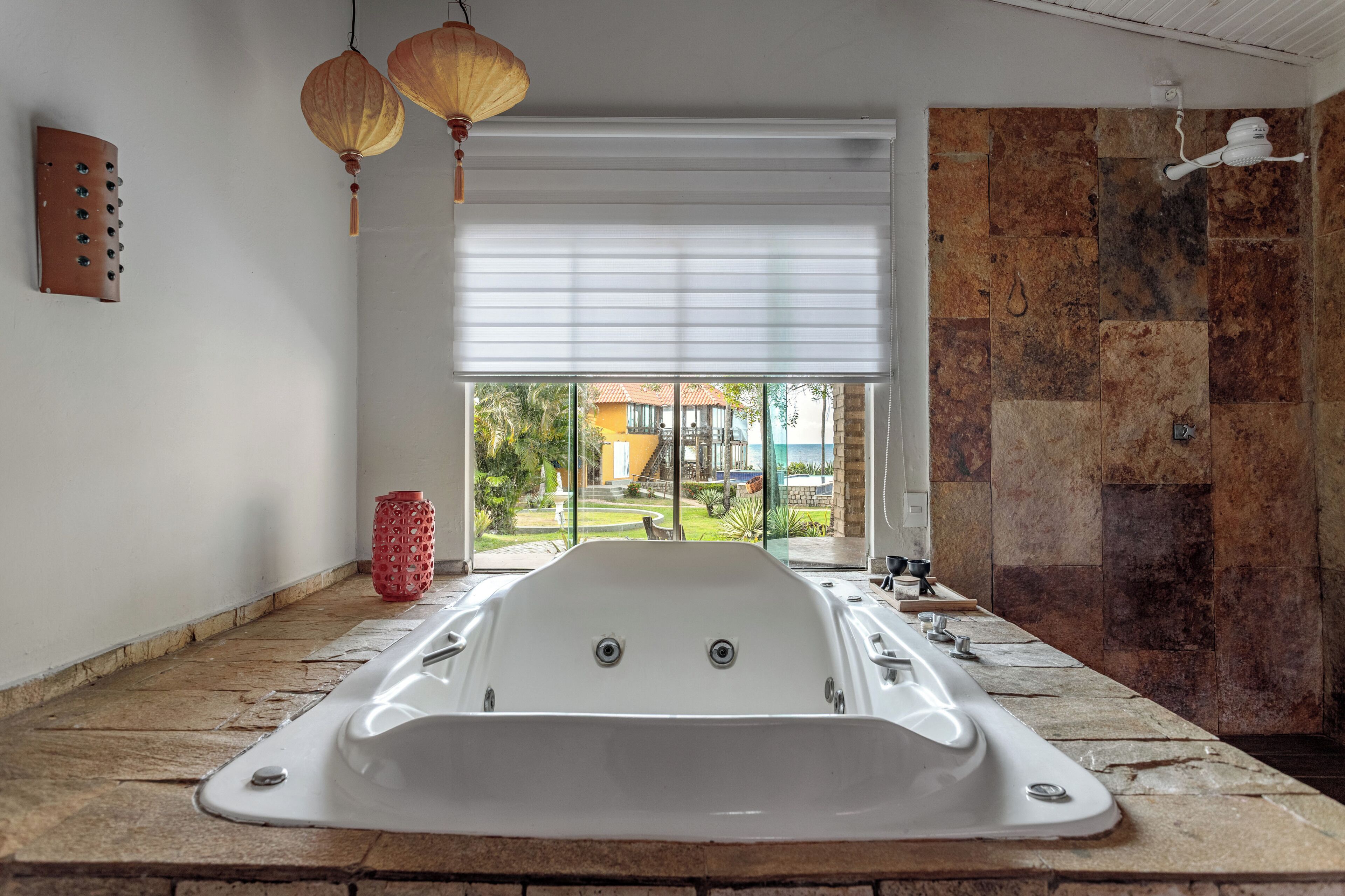 A modern bathroom with a large bathtub, a shower area, and a view of a garden and building through a large window.