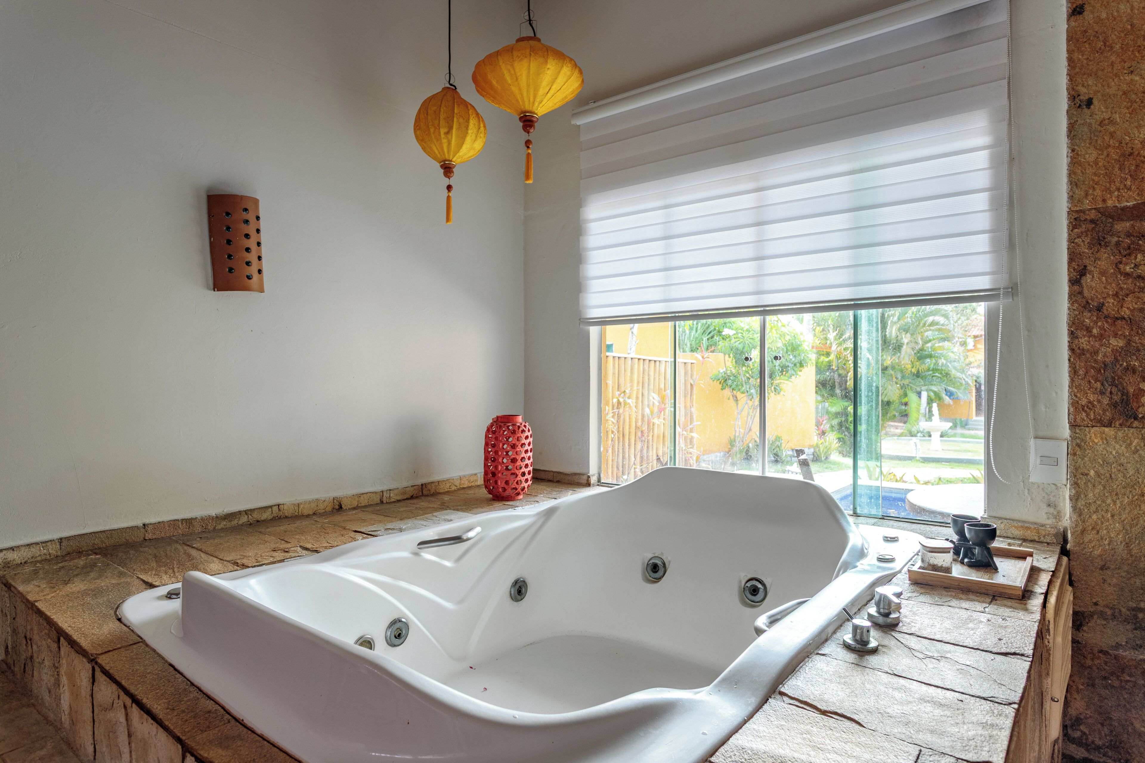 Un baño moderno con una bañera independiente, un ventanal con persianas y un florero decorativo rojo.