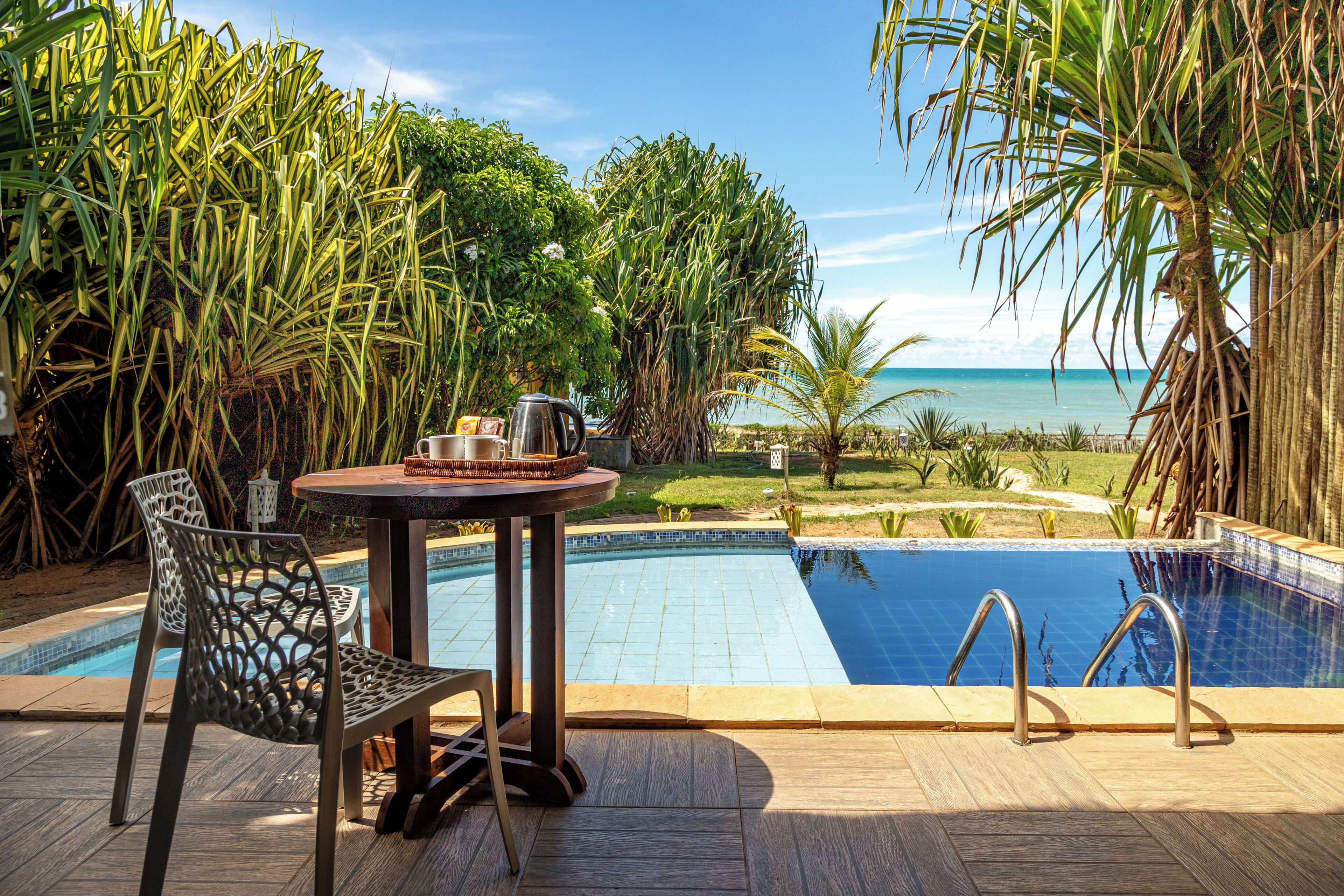 Ein Poolbereich mit Tisch und Stühlen, Blick auf den Ozean und üppiger tropischer Vegetation.