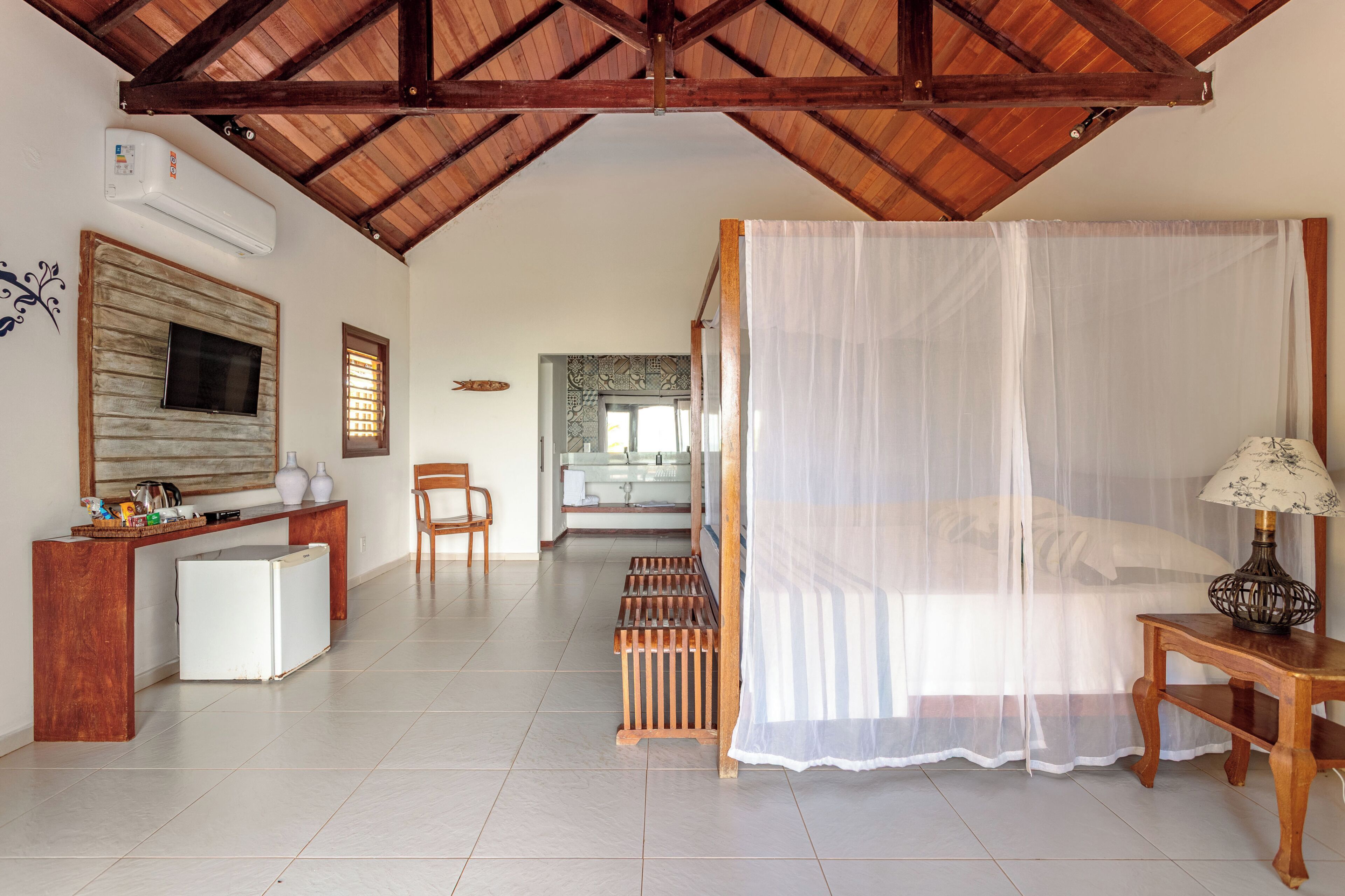A bedroom with a canopy bed, a desk with a lamp, a TV mounted on a wooden panel, a small refrigerator, and a bathroom visible in the background.