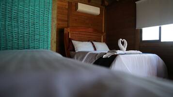 A room with two beds, wooden walls, a window with a white blind, and a woven wall hanging.