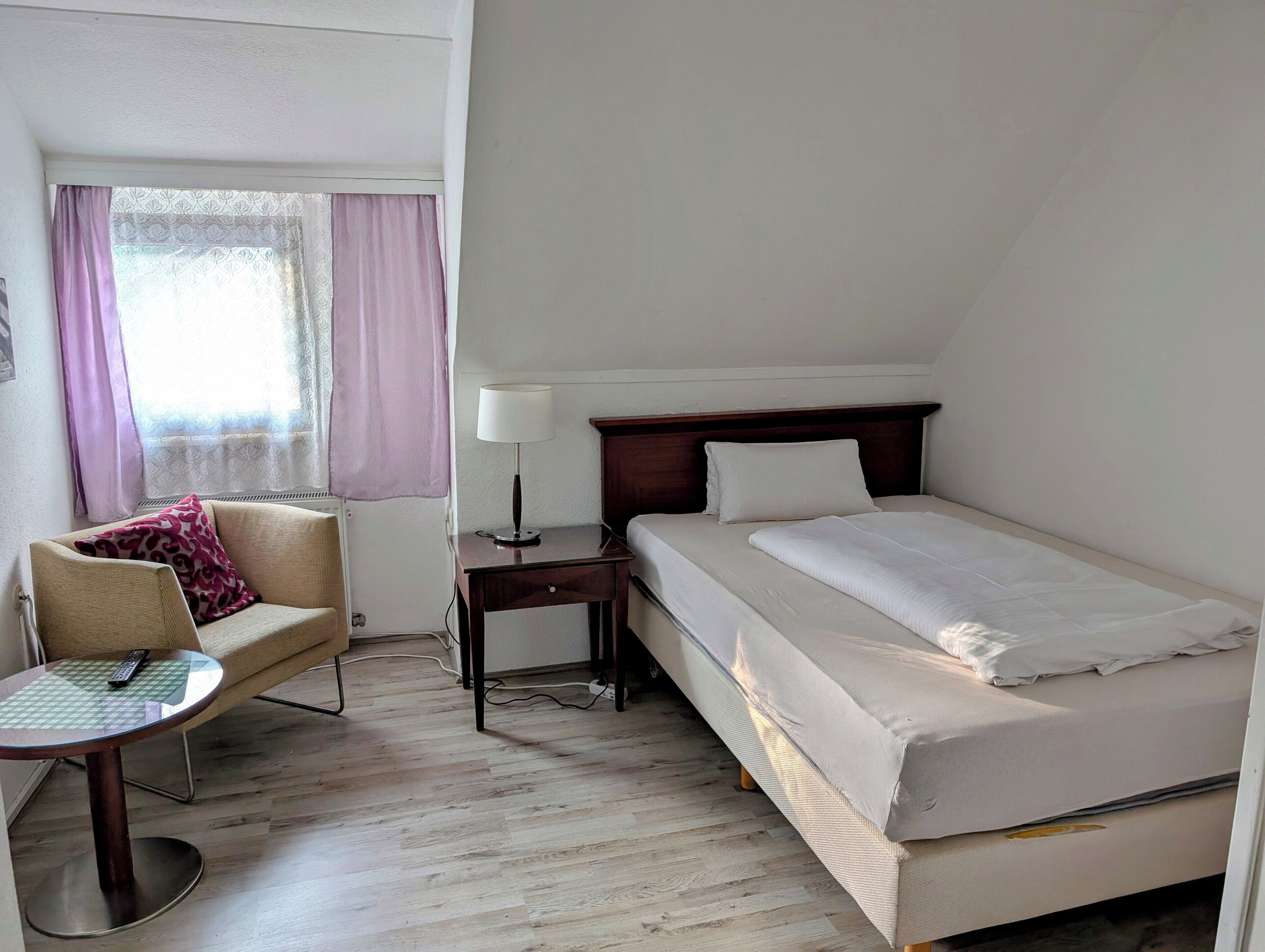 A hotel room with a bed, a chair, a small table, and a lamp.
