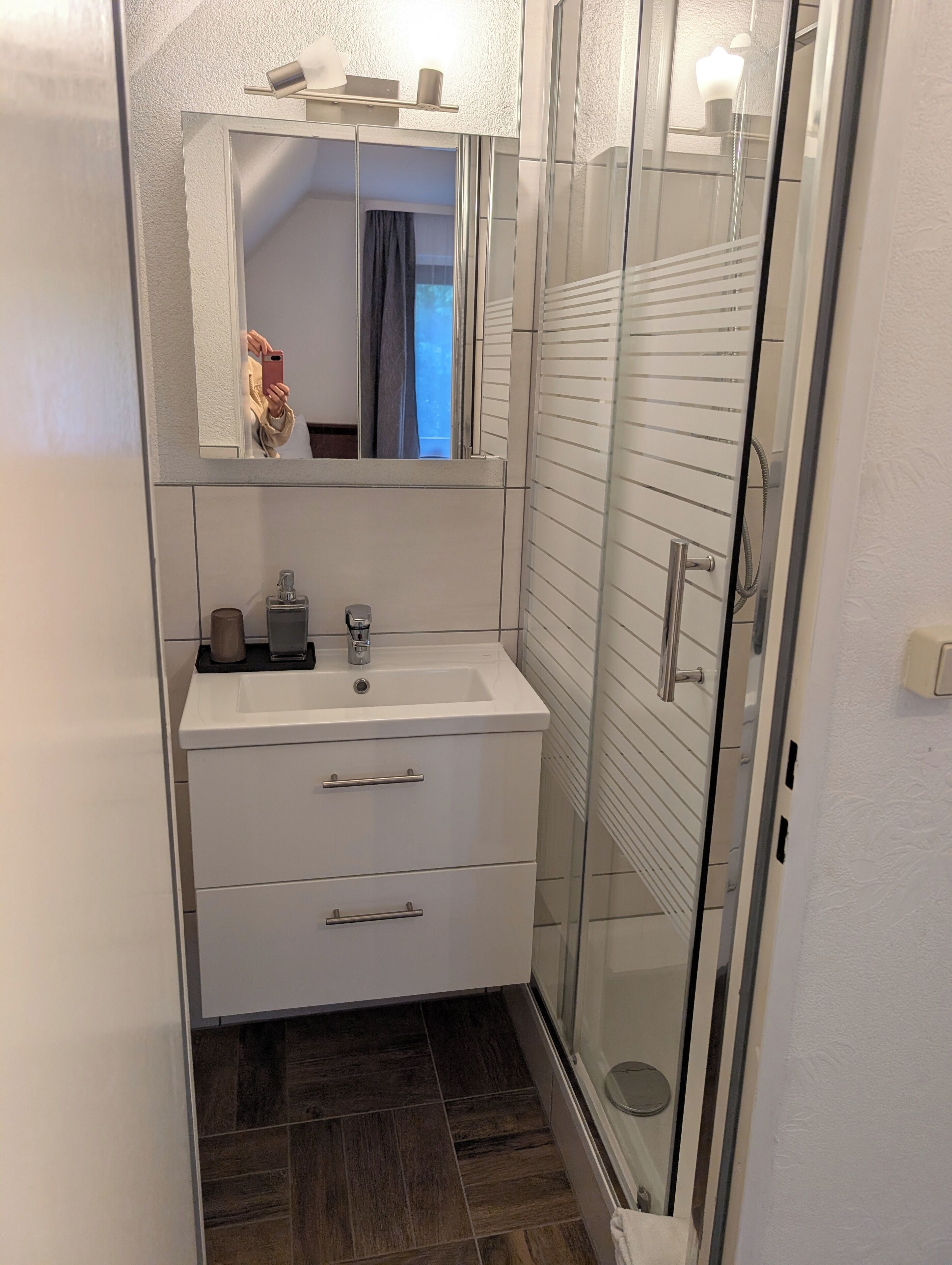 A modern bathroom with a glass shower enclosure, a white sink with a mirror above it, and a dark wood floor.