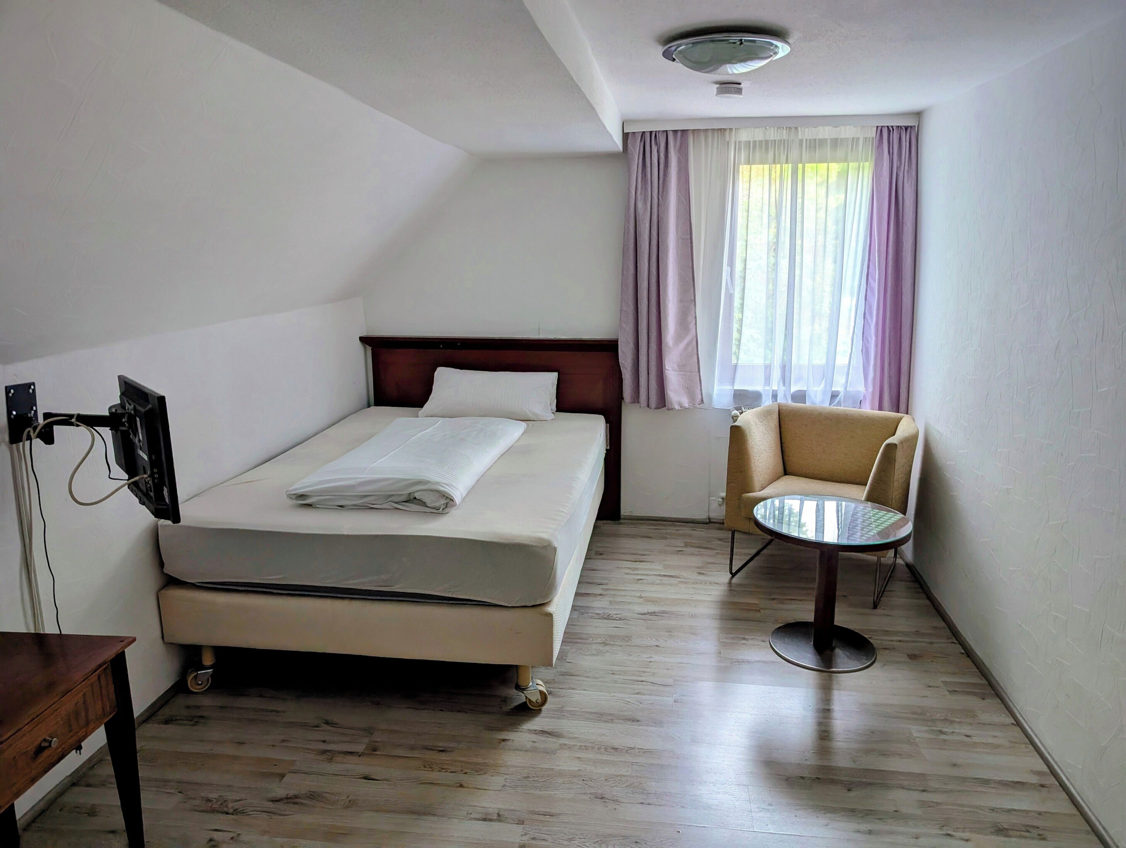 Et hotelværelse med seng, stol, lille bord og et fjernsyn ophængt på væggen.