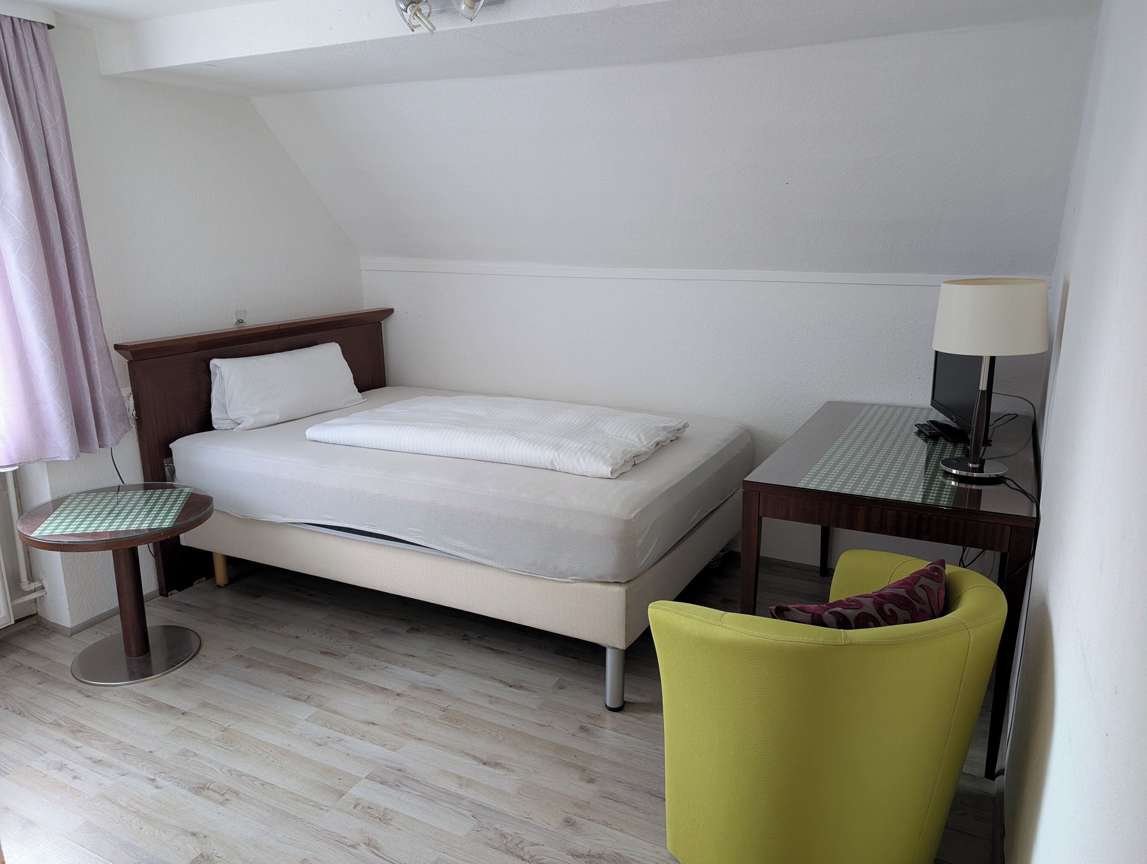 A hotel room with a bed, a small table, a chair, a desk with a lamp, and a window with curtains.