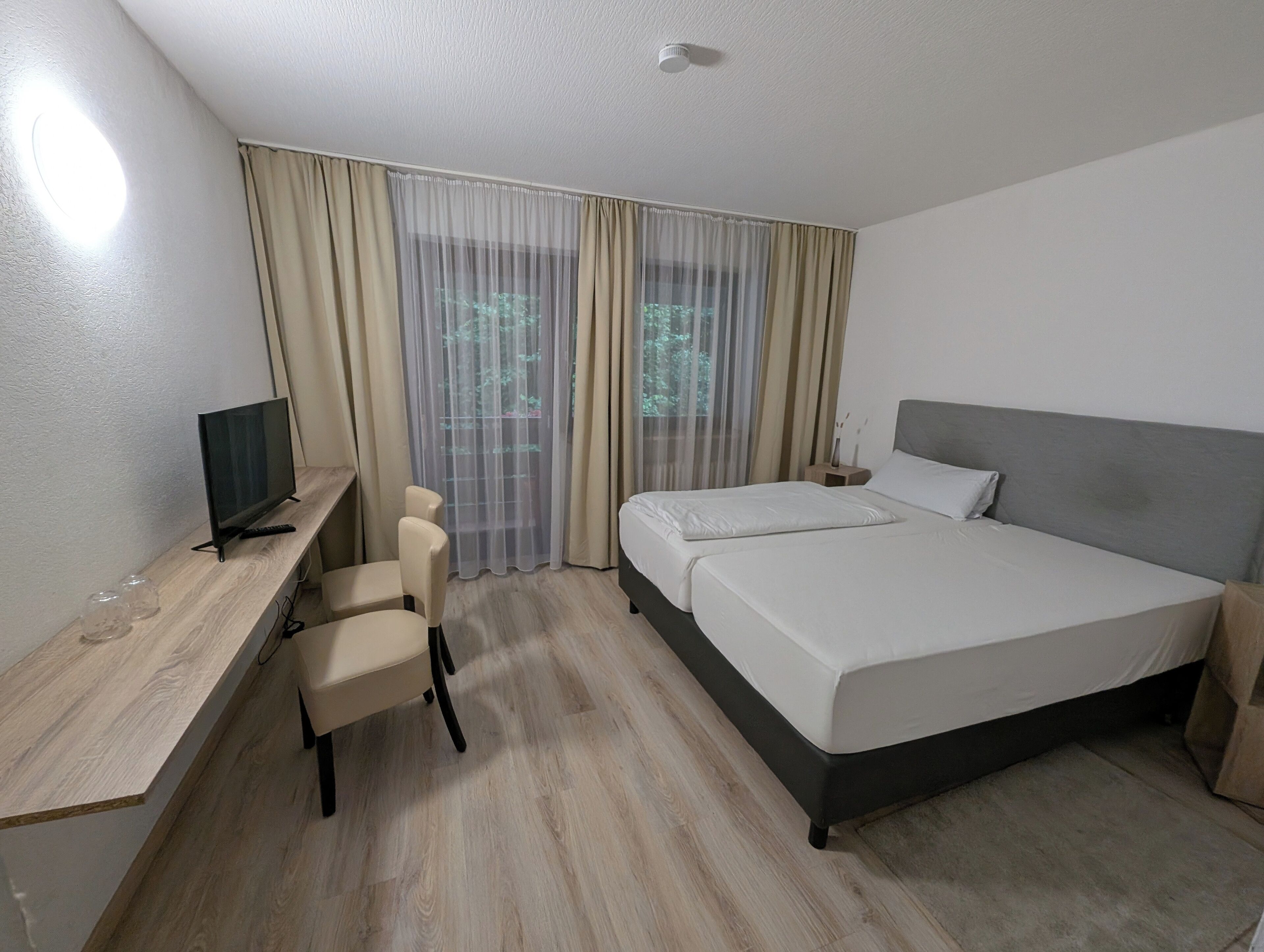 A hotel room with a bed, a desk with a TV, a chair, and a window with curtains.