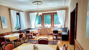 Comedor con varias mesas y sillas, ventanas amplias con cortinas y vistas a la vegetación exterior.