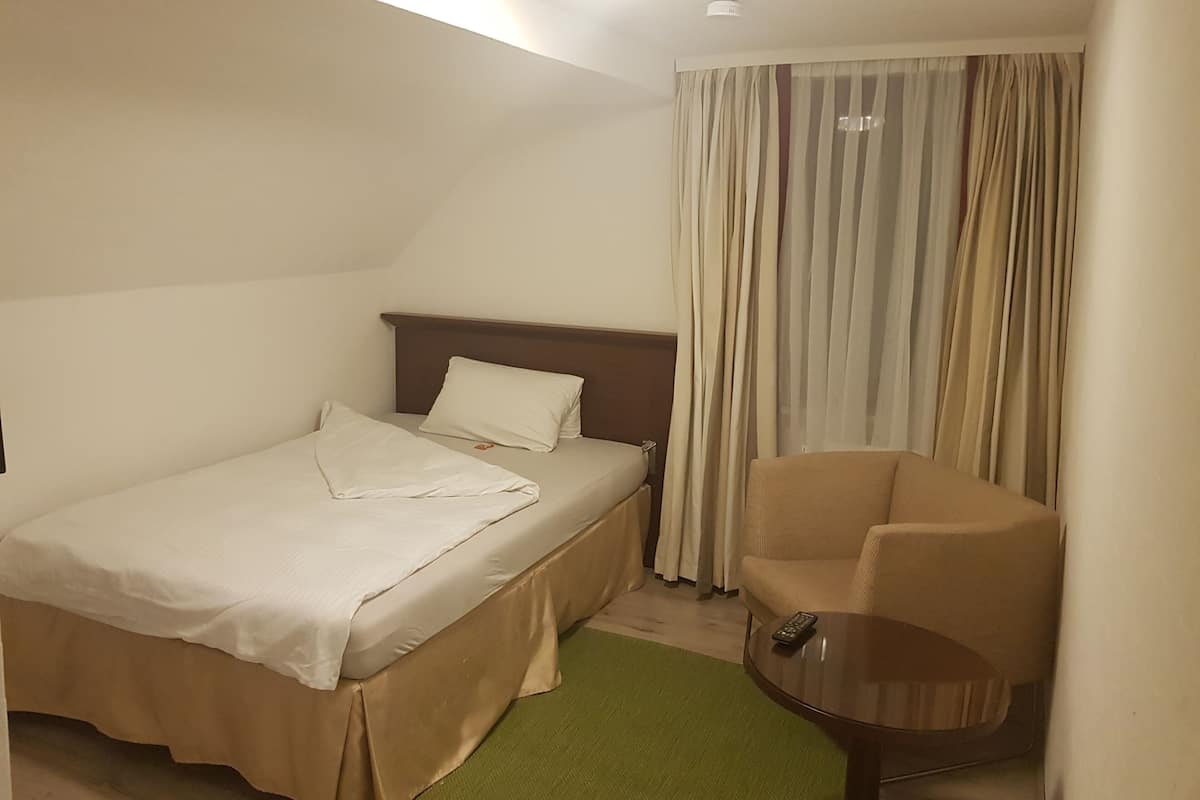 A hotel room with a single bed, a chair, a small table, and a window with curtains.
