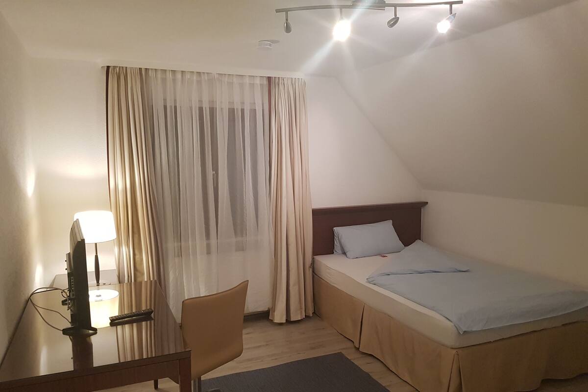 A small, well-lit room with a single bed, a desk with a chair, and a TV.