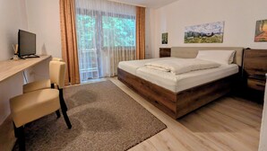 Un dormitorio con una cama de madera, un escritorio con una computadora, una silla y vista a zonas verdes por la ventana.