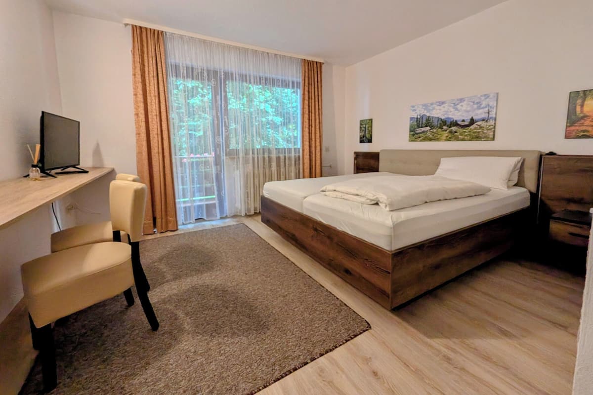 A bedroom with a wooden bed, a desk with a computer, a chair, and a view of greenery through the window.