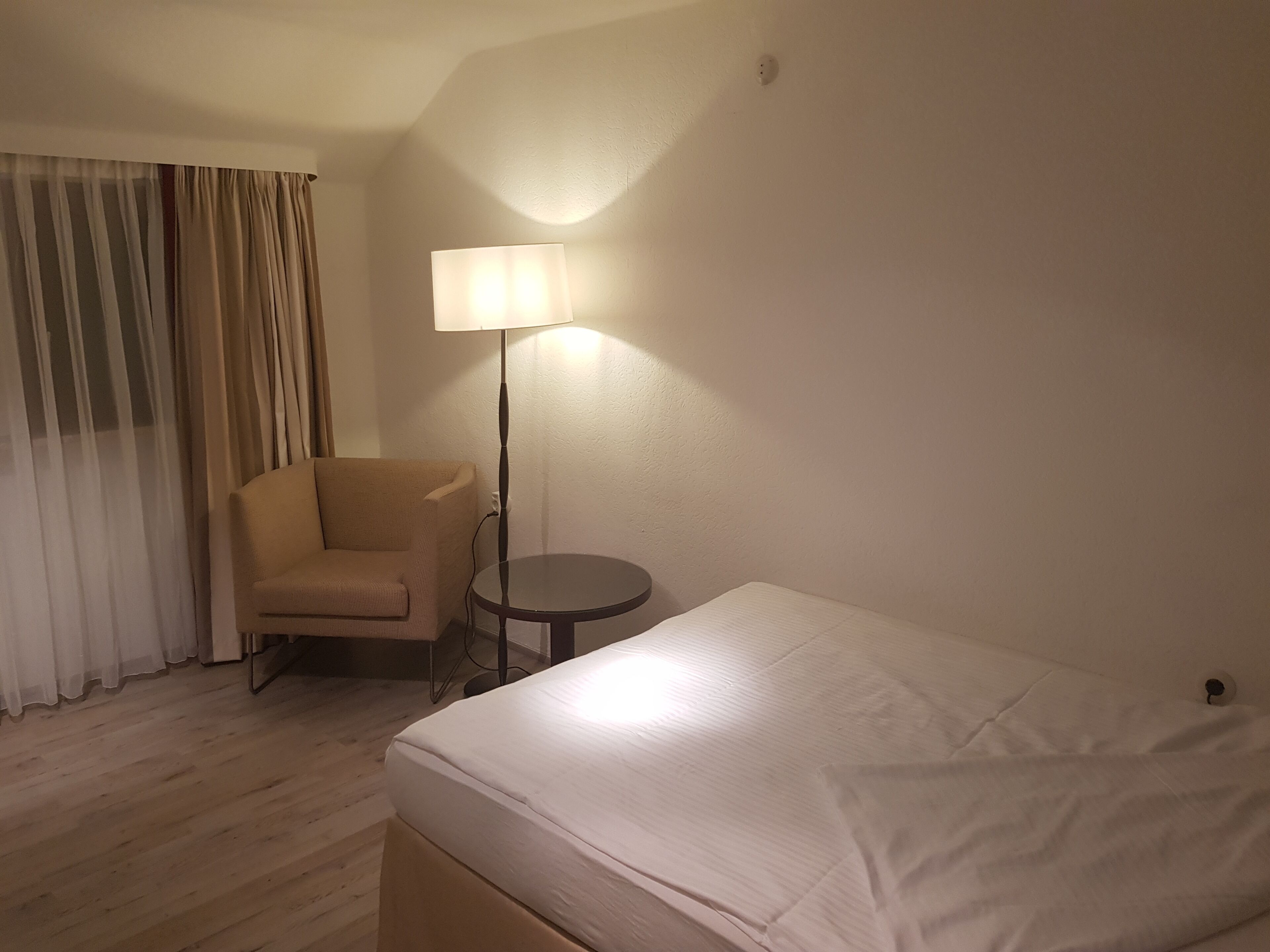 A hotel room with a bed, a chair, a small table, and a lamp.