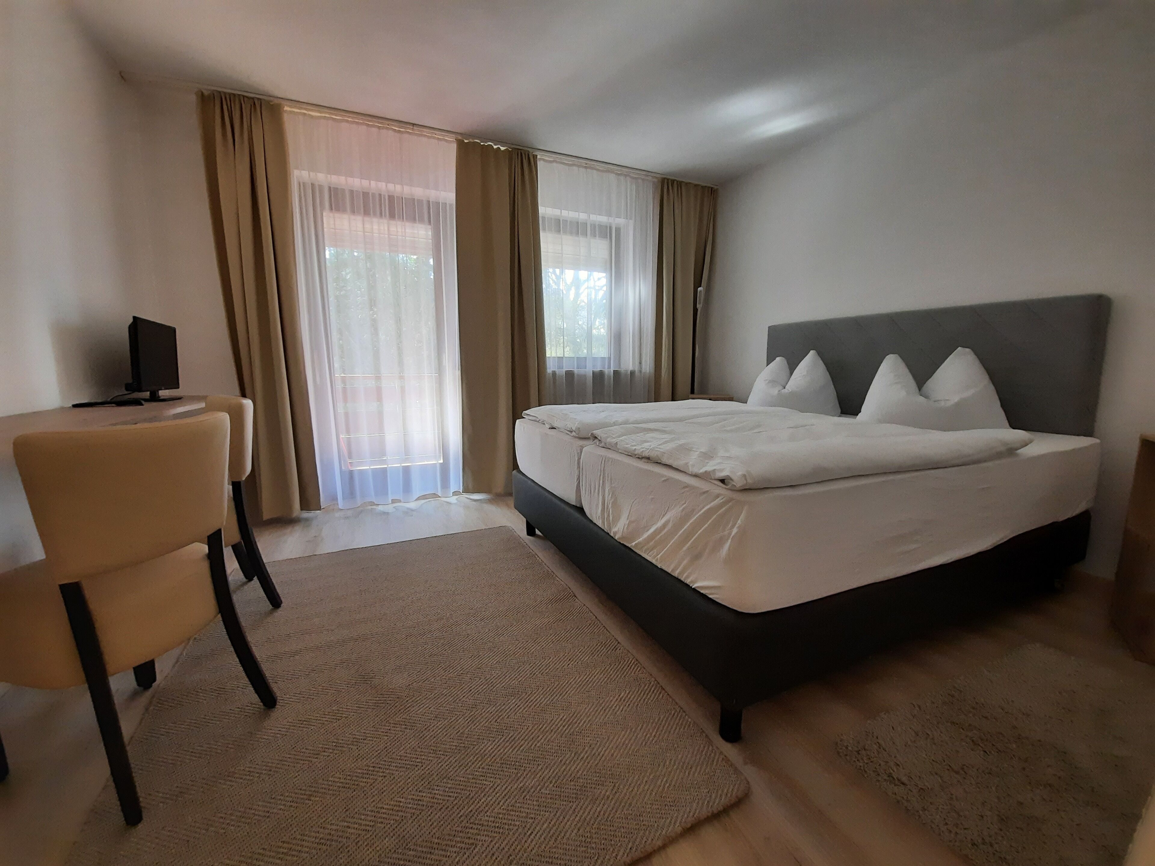 A bedroom with a bed, a chair, a desk with a computer, and a window with curtains.