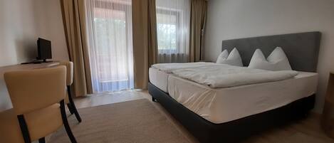 A bedroom with a bed, a chair, a desk with a computer, and a window with curtains.