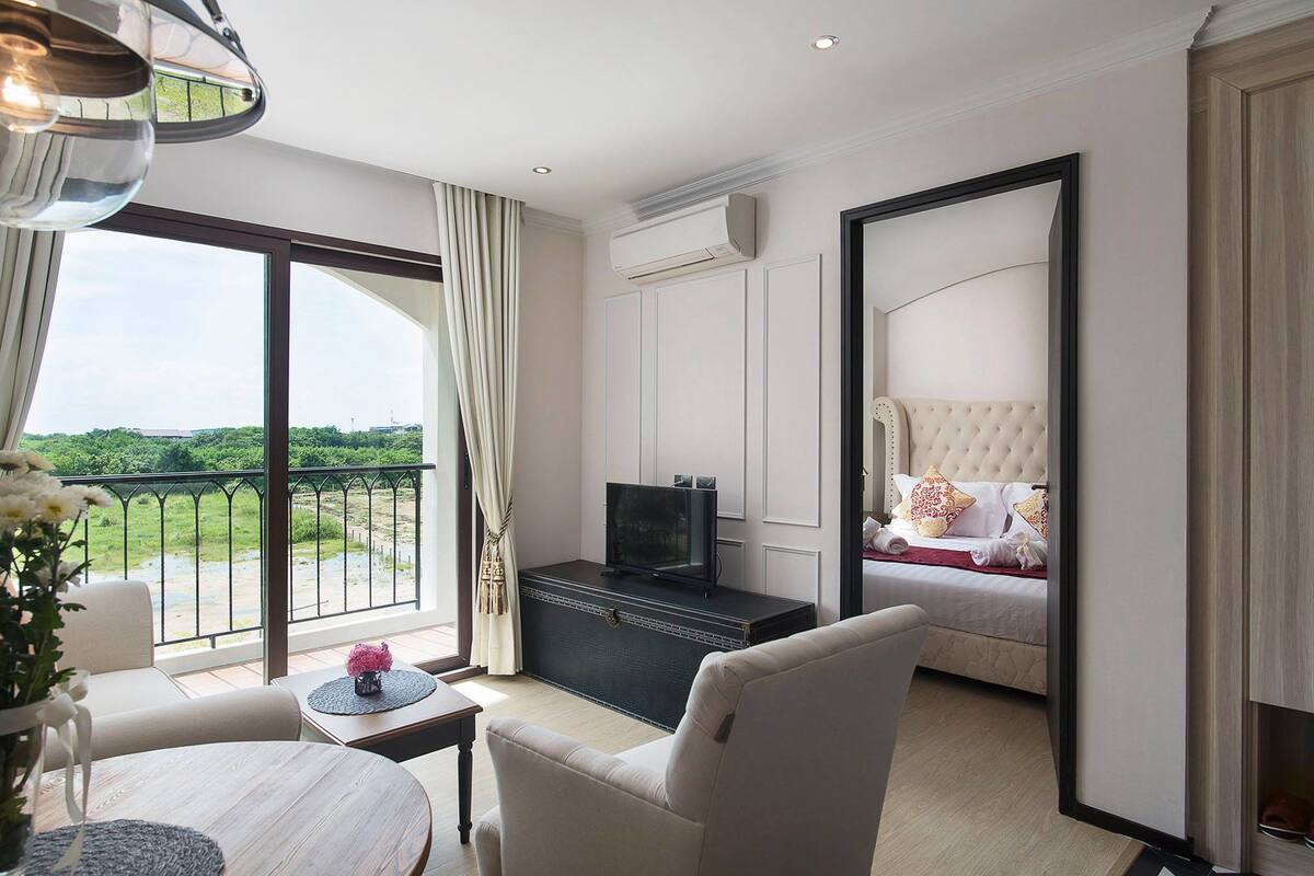 A hotel room with a balcony, a bed with a tufted headboard, a television on a dresser, and a small table with a vase of flowers.